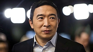 A man in a suit stands indoors with blurred lights and people in the background.