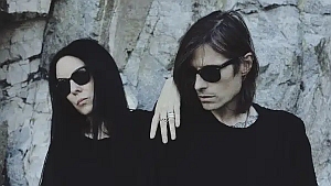 Two people wearing black clothes and sunglasses stand against a rocky background, with one person's arm resting on the other's shoulder.