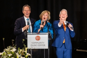 Three people in formal attire sing into microphones behind a podium labeled "Community Music Center," with a floral arrangement in front.