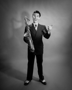 Man in a suit standing and holding a saxophone in one hand and a cigar in the other, smiling at the camera in a studio setting.
