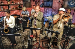 A jazz band performs indoors, with musicians playing saxophone, guitar, trumpet, trombone, and drums. Shelves and brick walls are visible in the background.