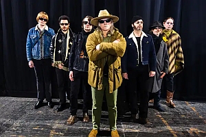 Seven people stand indoors on a stage in front of a black curtain, wearing sunglasses and various hats and jackets, with one person posed in front with arms crossed.