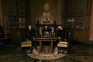 Three men sit around a wooden table in a dimly lit, ornate library with bookshelves, large chairs, and a painting above a fireplace.