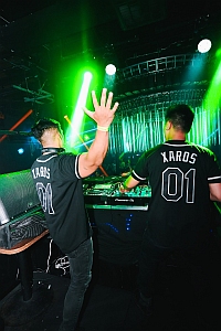 Two DJs wearing matching "KAROS 01" shirts perform at a nightclub, illuminated by green stage lights and lasers, with equipment in front of them.