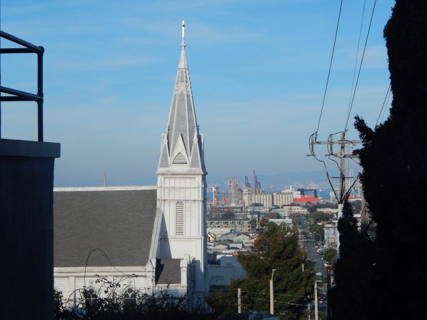 A white church steeple with a cross stands in the foreground, overlooking an urban area with industrial buildings and cranes under a clear sky.