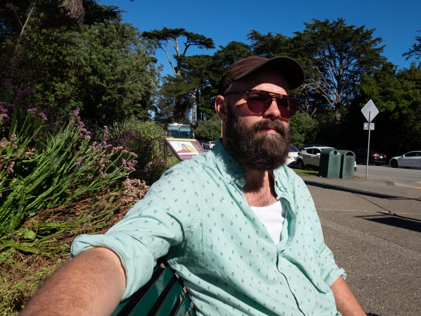 A bearded man wearing sunglasses, a cap, and a light green shirt sits on a bench outdoors on a sunny day, with trees and parked cars in the background.