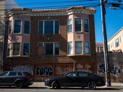 A two-story brick and stucco apartment building with boarded-up windows and graffiti on the ground floor; two parked cars are in front.