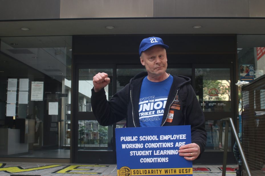 A man in a blue cap and shirt stands outside a building, raising his fist and holding a sign supporting teacher and public school employee working conditions.