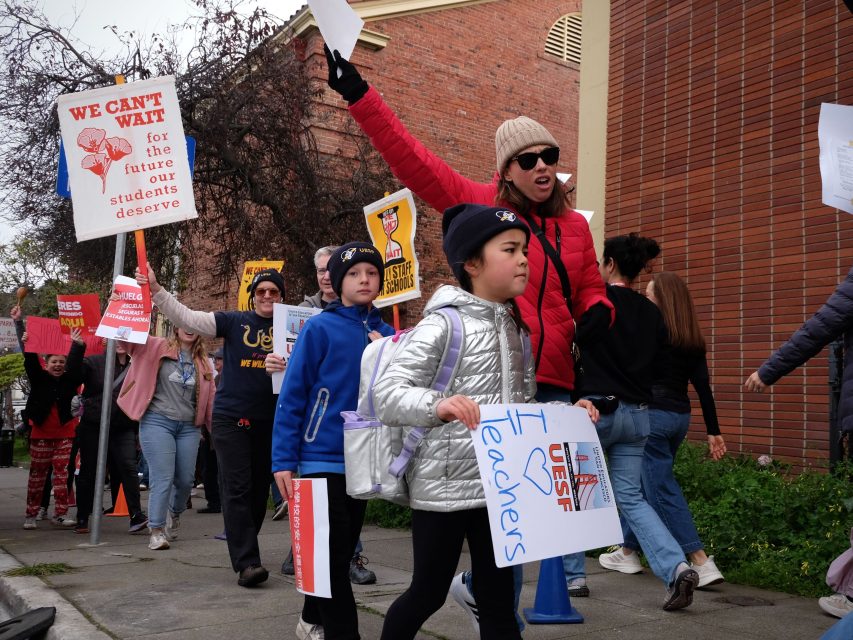 A group of people, including children, march on a sidewalk holding signs in support of teachers and education.