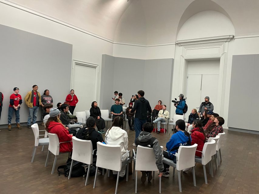 A group of people, some seated in a circle and others standing, gather in a spacious room with white walls as a person speaks in the center; a camera crew is present.