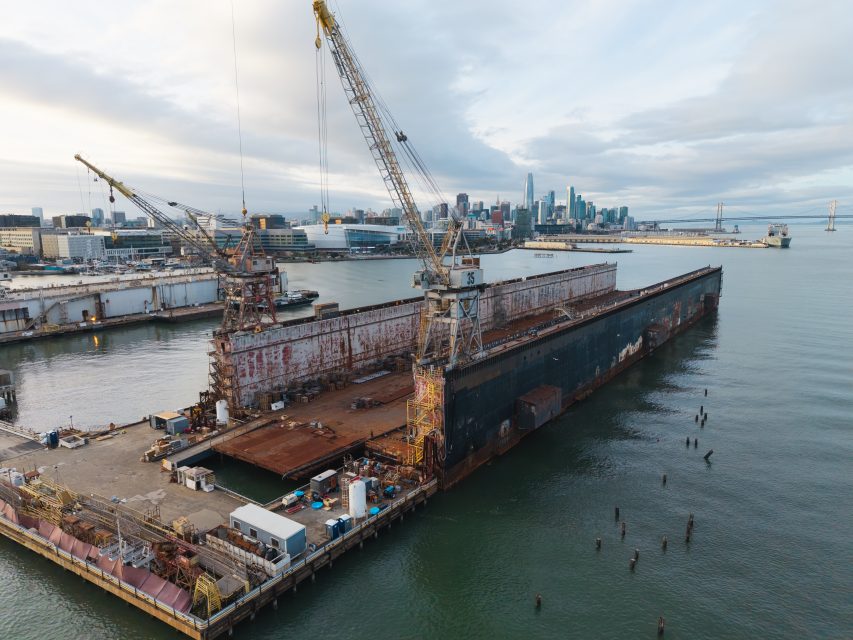 Large industrial cranes stand on a floating dry dock in a harbor, with a city skyline and bridge visible in the background under a cloudy sky.