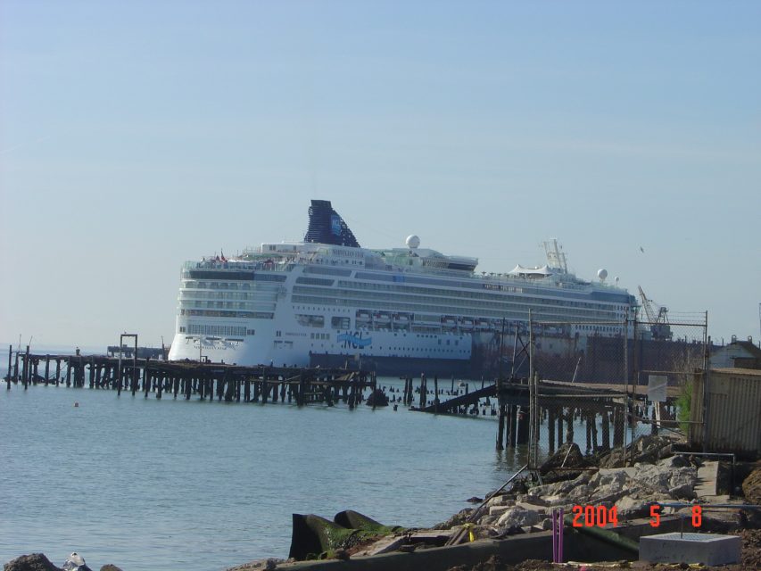 A large white cruise ship is docked near a weathered pier beside rocky shoreline under a clear sky. The date on the image reads 2004-05-08.