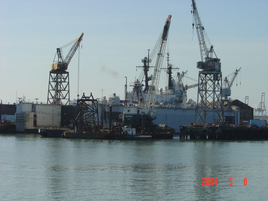 Industrial shipyard with cranes, docked ships, and barges seen from across the water under a clear sky. The date stamp reads 2004-05-08.