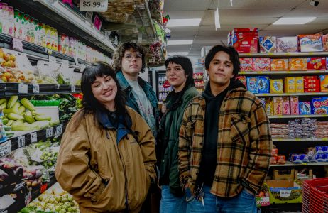 Four people stand together in a grocery store aisle near produce and packaged goods, looking at the camera.