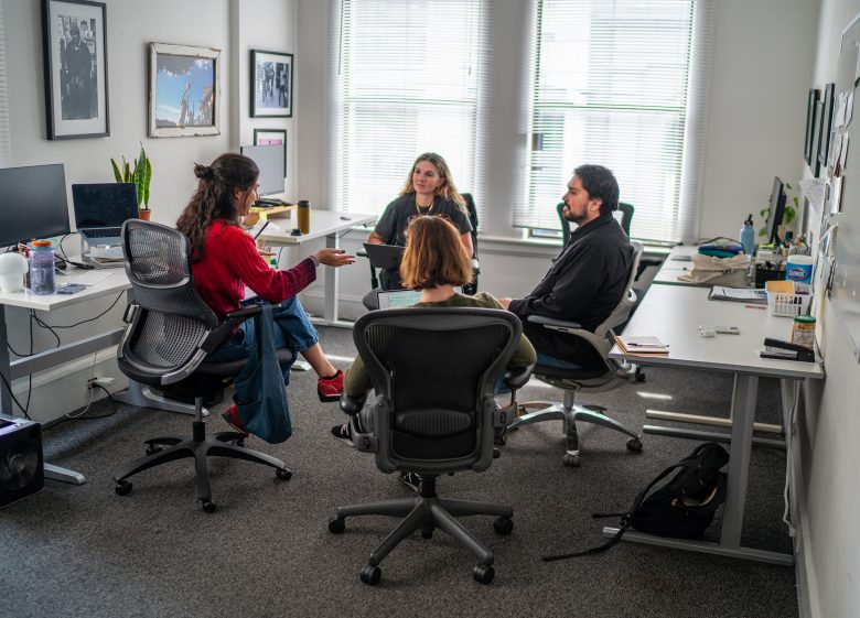 Four people sit in office chairs in a brightly lit office, engaged in a discussion around a group of desks with computers and office supplies.