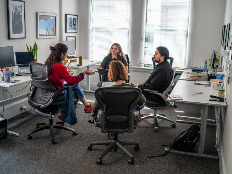 Four people sit in office chairs in a brightly lit office, engaged in a discussion around a group of desks with computers and office supplies.