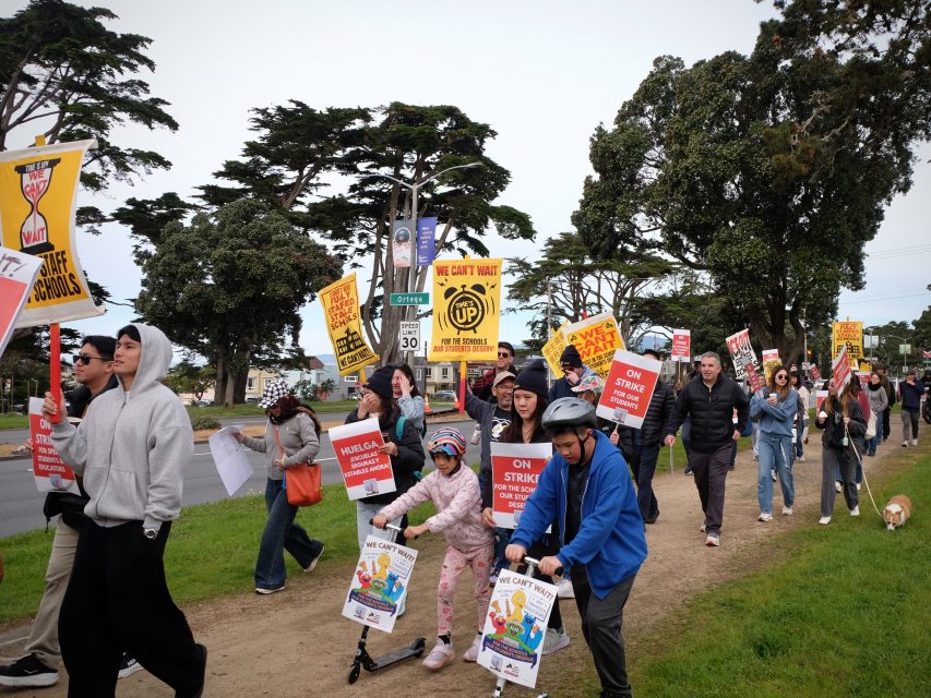 A group of people march outdoors holding signs that read “On Strike,” “We Can’t Wait,” and “We Love Our Schools,” with trees and street signs in the background.