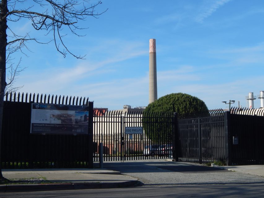 A tall industrial smokestack stands behind a black metal fence and gate, with a bare tree on the left and informational signage visible near the entrance.
