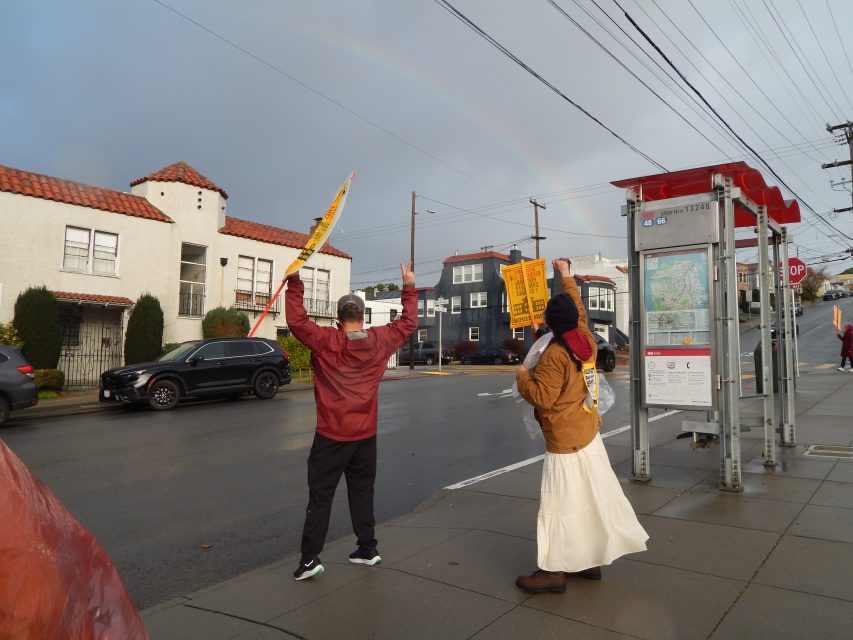 Two people stand on a sidewalk holding teachers strike protest signs near a bus stop on a cloudy day, with a faint rainbow visible in the sky.