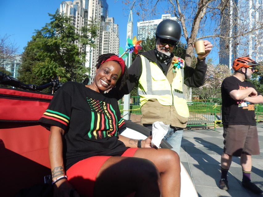 A woman sits and smiles at the camera while two men stand nearby; all are outdoors in an urban park with tall buildings in the background.