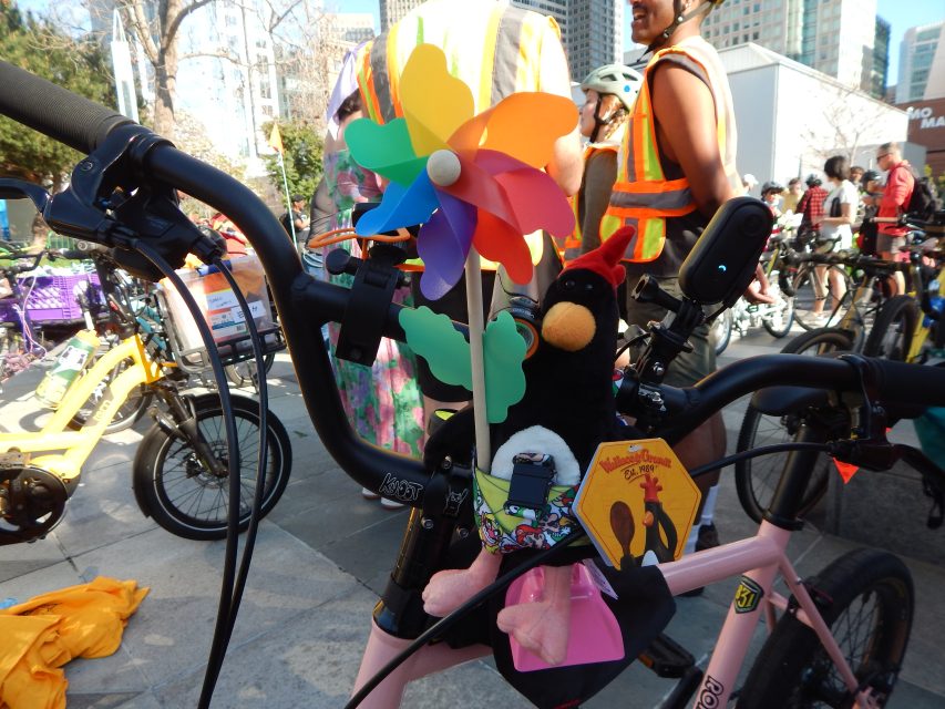 Close-up of a bicycle handlebar decorated with a colorful pinwheel, a black plush toy, and various accessories at a group cycling event. People and other bikes are in the background.