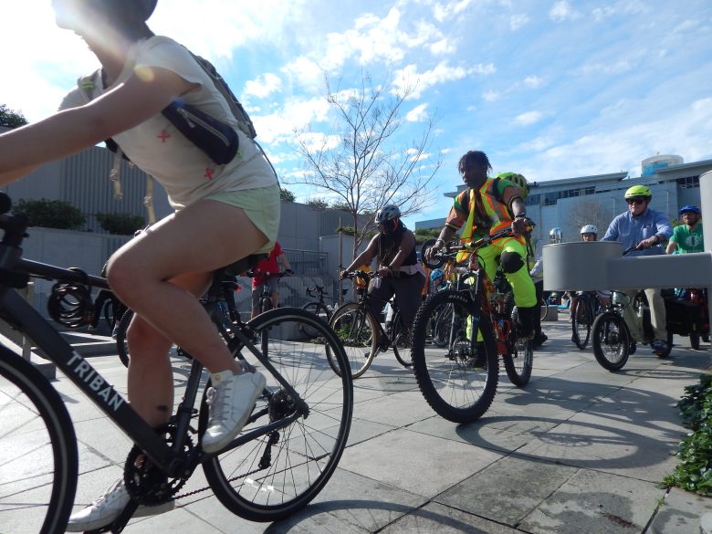 A group of cyclists, including people in casual and high-visibility clothing, ride together on a sunny day through the urban streets of Bayview.