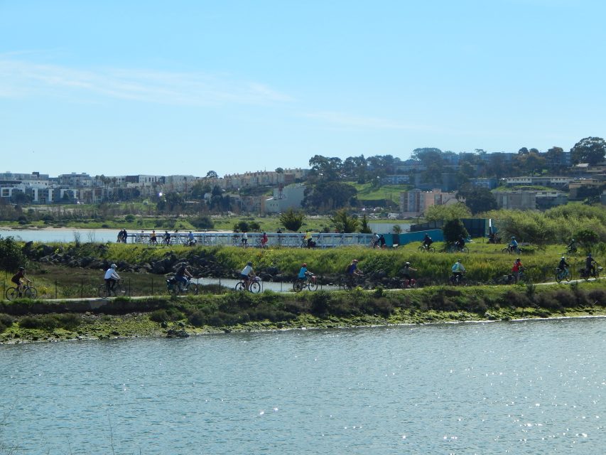 People ride bicycles along a path beside a body of water, with green vegetation and buildings visible in the background under a clear sky.