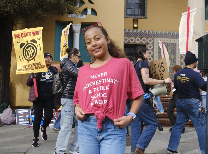 A girl stands confidently at a school protest wearing a shirt that says "Never underestimate a public school teacher," joining fellow teacher supporters holding signs in the background.