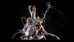 Five dancers in light gray costumes pose dramatically against a black background, with water splashing around them, suggesting motion and energy.