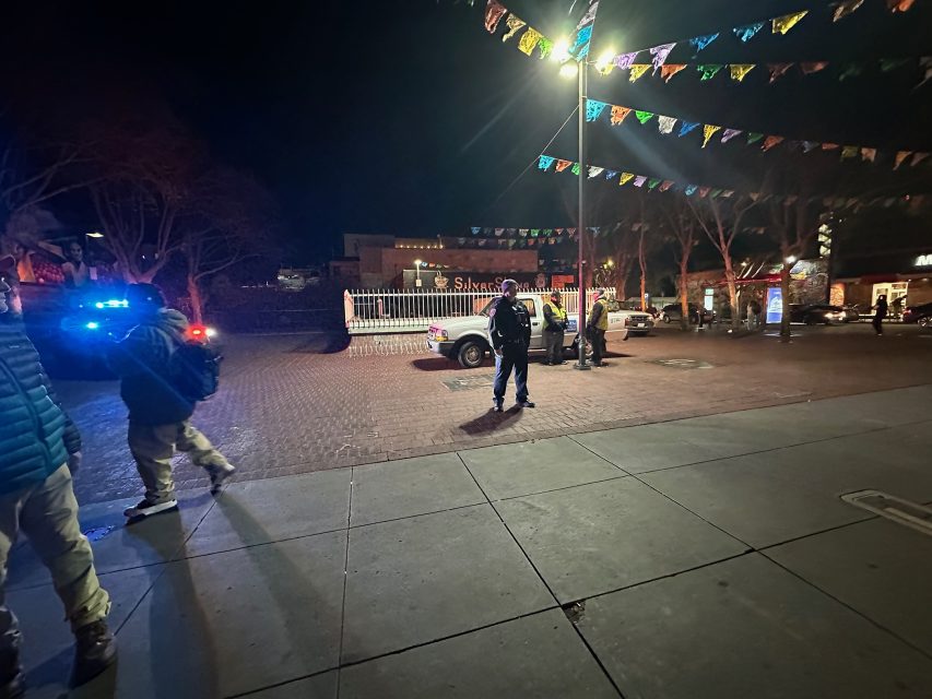 Police officers stand near patrol cars with flashing lights at night in a plaza decorated with colorful hanging banners; several pedestrians are nearby.