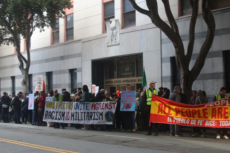 A group of people protest outside a building with banners reading "Our Faiths Unite Against Racism & Militarism" and "All People Are Equal.