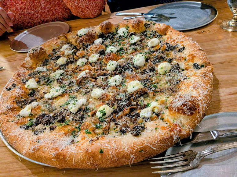 Large pizza topped with mushrooms, cheese, herbs, and grated parmesan on a wooden table with cutlery, plates, and people sitting nearby.