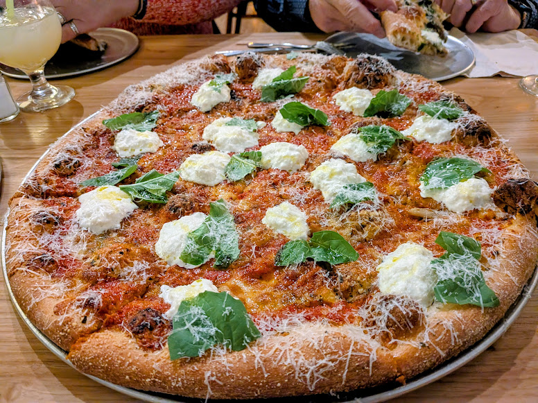 Large pizza topped with dollops of ricotta cheese, fresh basil leaves, grated parmesan, and tomato sauce on a wooden table in a restaurant setting.