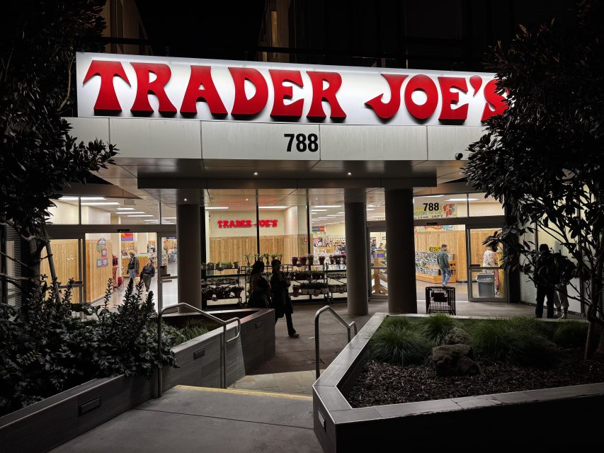 Exterior view of a Trader Joe's store at night, with illuminated signage above the entrance and a few people entering or exiting—perhaps picking up last-minute Valentine's Day treats.