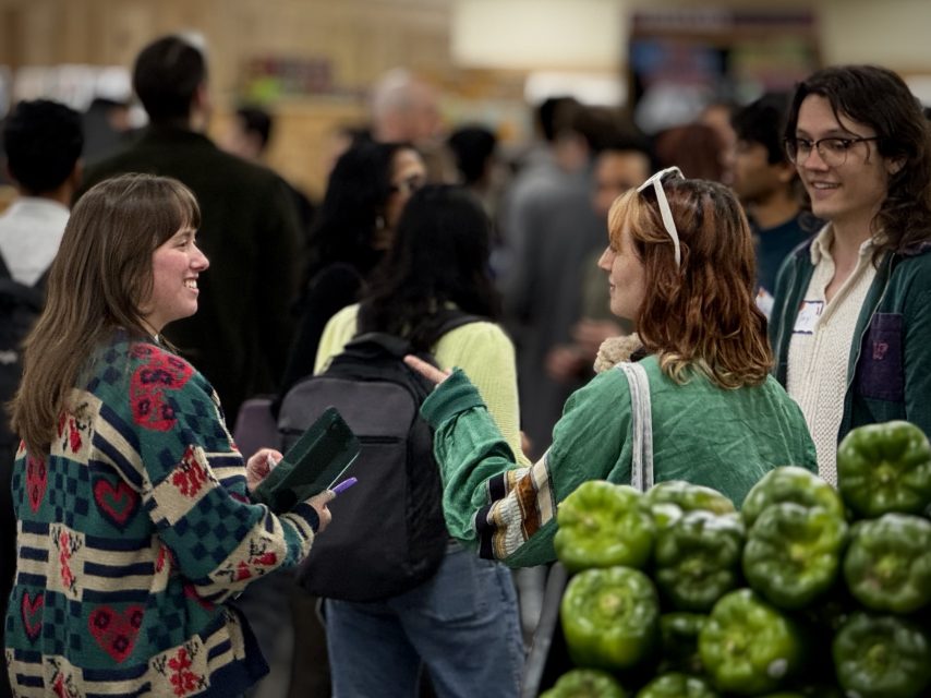 Three people stand and talk near a display of green bell peppers in a crowded indoor market, perhaps selecting fresh ingredients for a special Valentine's Day meal.