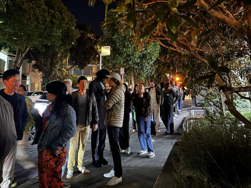 A line of people standing on a sidewalk at night, illuminated by streetlights, with trees and parked cars nearby, perhaps waiting to celebrate Valentine's Day together.