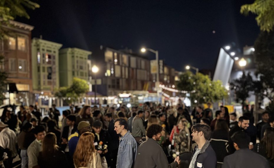 A large group of people gather outdoors at night on a city street, socializing under streetlights and string lights for Valentine's Day, with buildings and trees in the background.