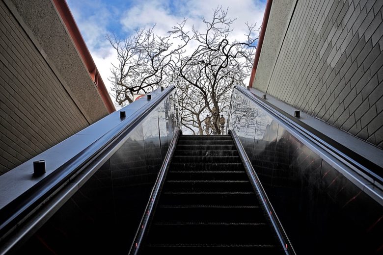 View from the base of an outdoor escalator looking up toward leafless tree branches and a partly cloudy sky between two building walls.