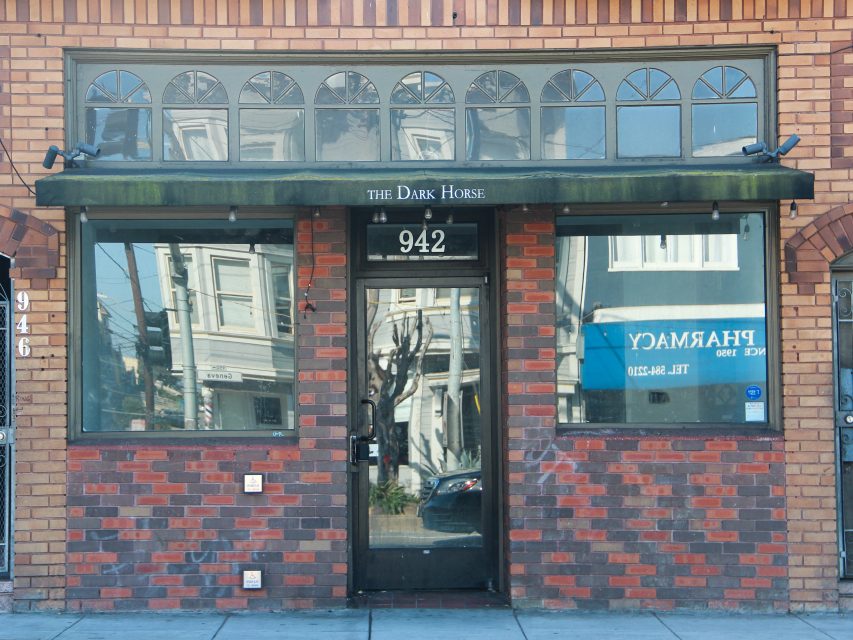 Storefront with red and brown brick exterior, glass door labeled "942," and an awning reading "THE DARK HORSE." Reflections of buildings—plus a blue Excelsior pharmacy sign—are visible in the windows.