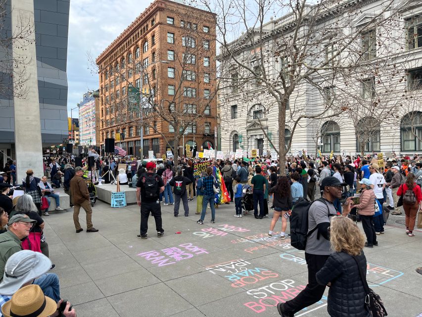 A large group of people gather for a protest in a city plaza, holding signs. Messages in chalk on the pavement include “STOP WAR NOW” and “STOP BOMBING.”.