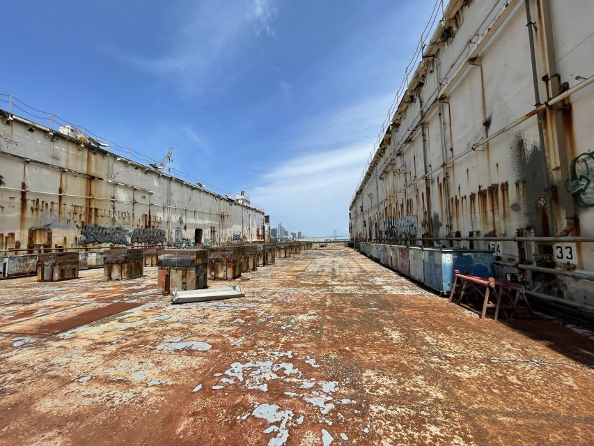 A wide, rusted, and weathered ship deck is flanked by two large, deteriorating structures with visible graffiti, under a partly cloudy blue sky.