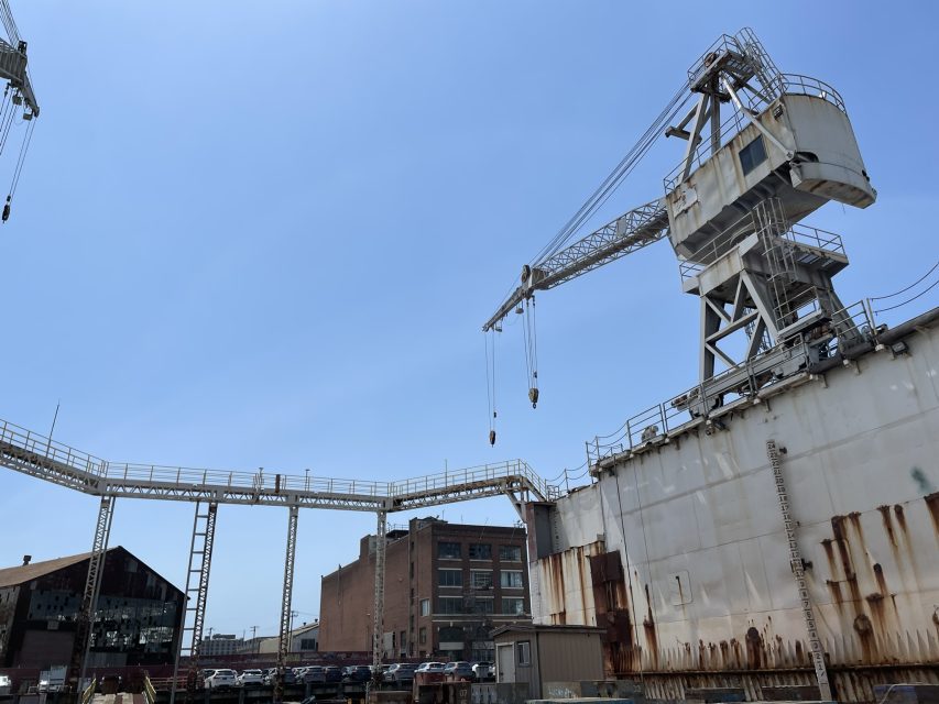Large industrial cranes and a weathered ship at a dockyard, with warehouses and parked cars in the background under a clear blue sky.