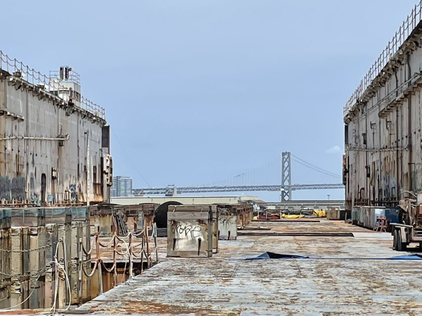 Two large, rusted industrial structures frame a view of the Bay Bridge in the distance under a clear sky.