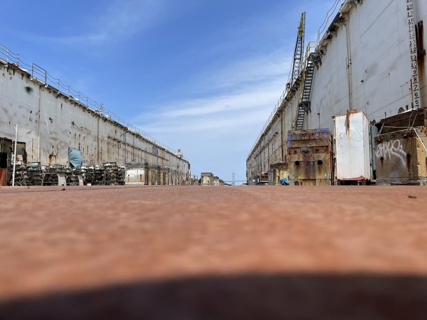 A low-angle view of an empty, rusted shipyard with large, worn metal walls on both sides under a blue sky.