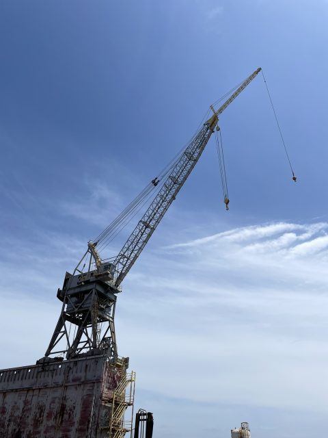 A large industrial crane extends upward against a blue sky with scattered clouds, situated on a weathered platform structure.