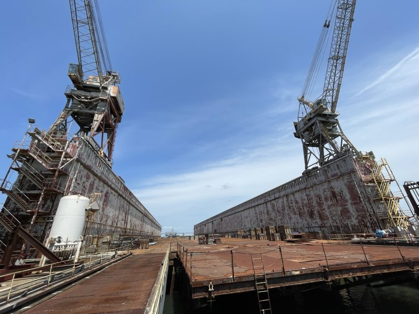 Two large industrial cranes stand on opposite sides of a rusty dry dock under a clear blue sky, with machinery and equipment scattered on the ground.