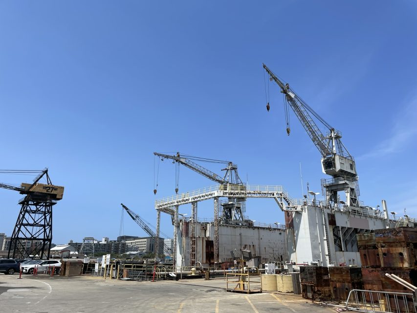 Large cranes and a docked ship at an industrial shipyard under a clear blue sky, with buildings visible in the background.