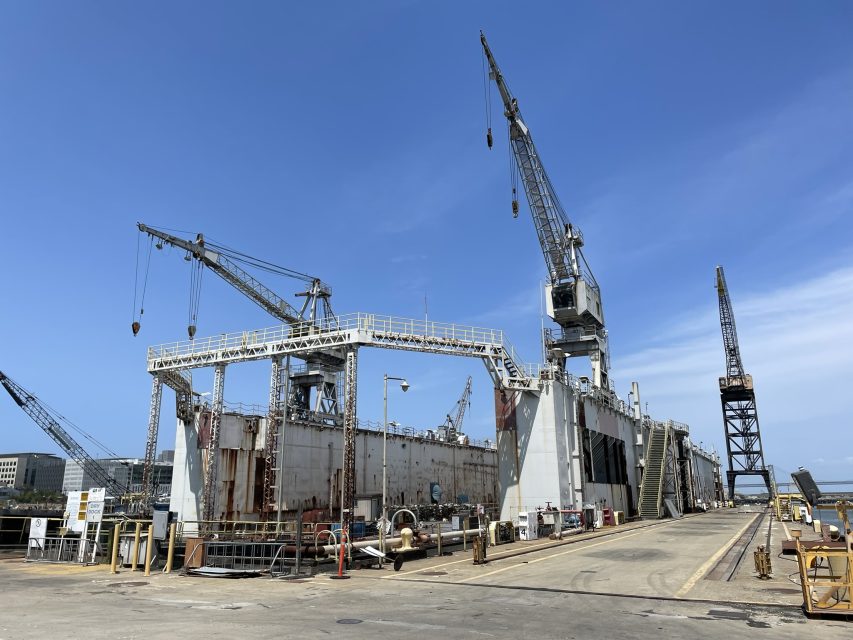 Large dry dock with multiple cranes and industrial equipment situated along a pier under a clear blue sky.