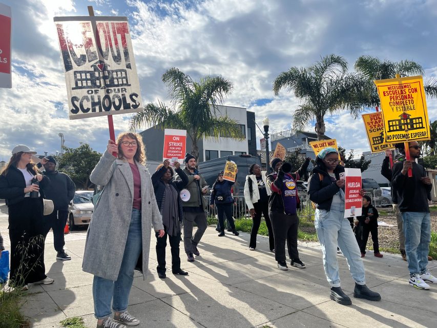 A group of people stand on a sidewalk holding signs advocating for schools and fair education, with palm trees and buildings in the background.