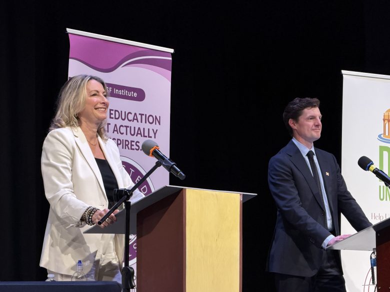 A woman and a man stand at separate podiums on a stage, each with a microphone, participating in what appears to be a public debate or discussion.
